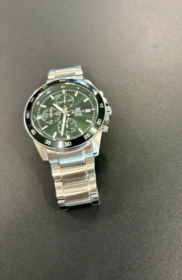 Zilver en groen analoog Casio horloge met datum aanduiding op grijze ondergrond.