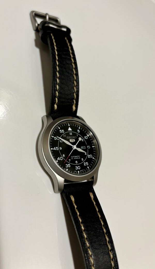 Zilver met zwart automatisch rond Seiko 5 horloge met witte wijzers and datum aanduiding met lederen band op witte ondergrond.