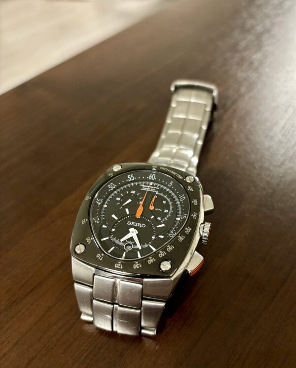 Zilver met zwart Seiko horloge met oranje wijzers en datum aanduiding op houten ondergrond.
